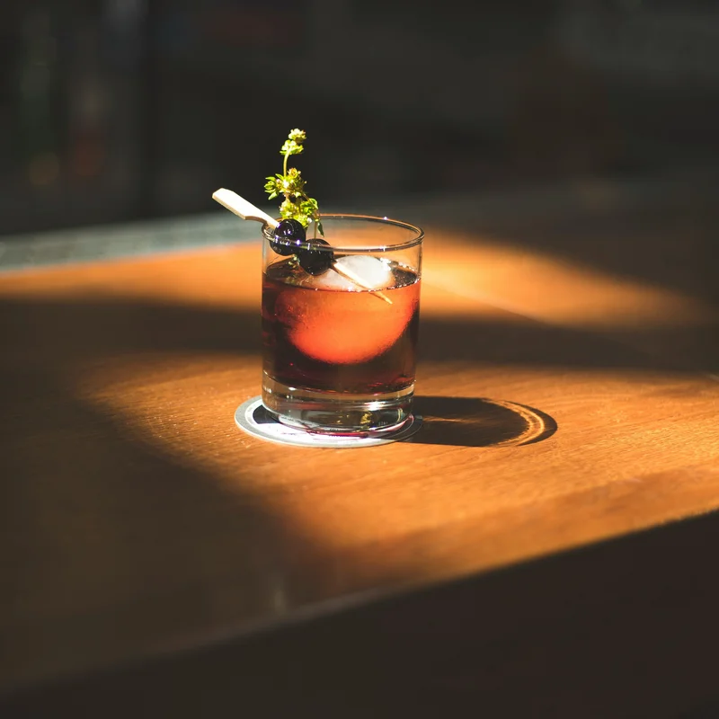 Was erwarten Sie von einer Bar in Ischgl? Cocktailglas mit braunem Getränk, Eiswürfel und Beeren auf Holzoberfläche