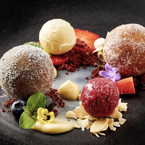 Ihr liebstes Restaurant in Ischgl Kunstvoll angerichtetes Dessert mit Kugeln, Beeren, Blüten und Creme auf schwarzem Teller