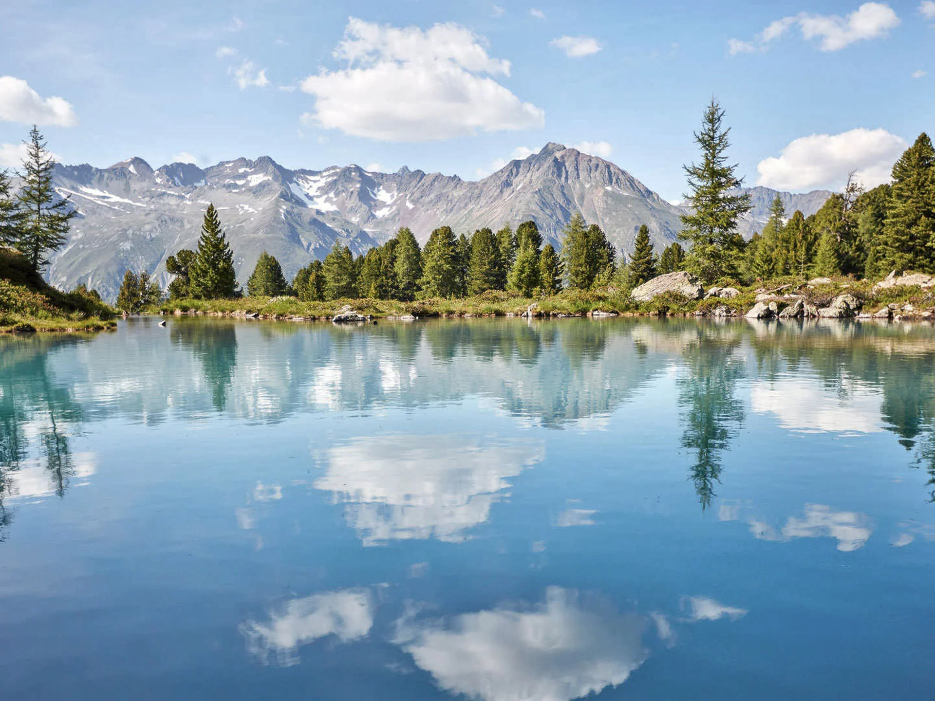 Pictures from Hotel Salnerhof Mountain lake reflecting trees, mountains, and clouds on a sunny day