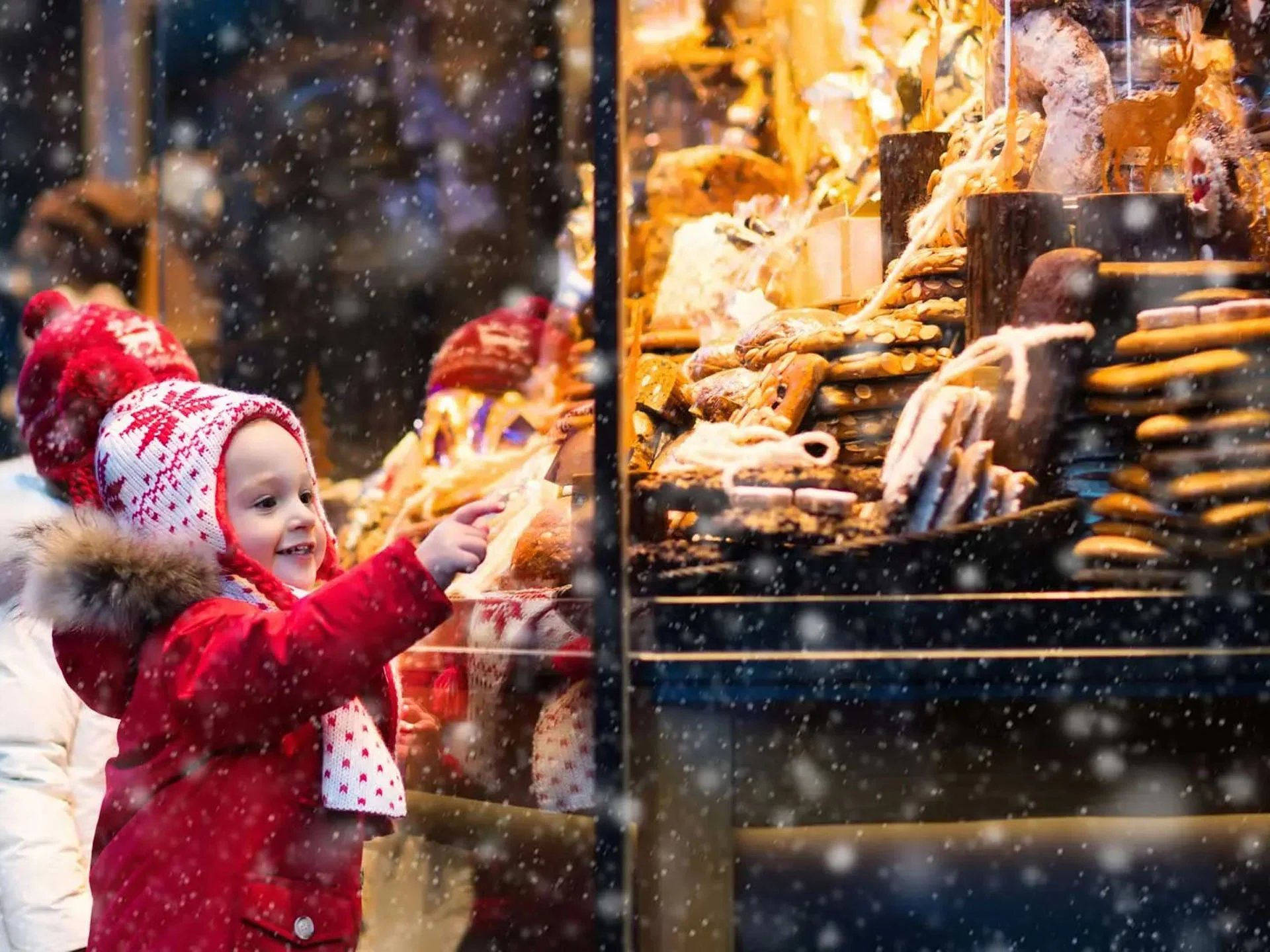Die Bilder aus dem Hotel Salnerhof Kind im roten Mantel zeigt auf Lebkuchen in einem weihnachtlichen Schaufenster bei Schneefall