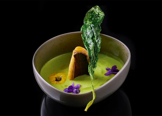 Ihr liebstes Restaurant in Ischgl Grüne cremige Suppe mit essbaren Blüten und knusprigem Blatt in weißer Schale