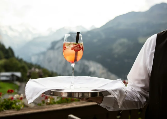 Kellner mit Inkasso Kellner hält ein Tablett mit Cocktailglas vor Bergpanorama
