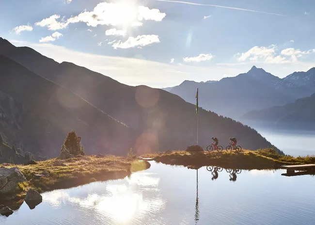 Ihr Sommer im Paznaun im Wander- und Bike-Hotel in Ischgl Zwei Radfahrer fahren entlang eines Bergsees bei Sonnenuntergang mit Bergen im Hintergrund