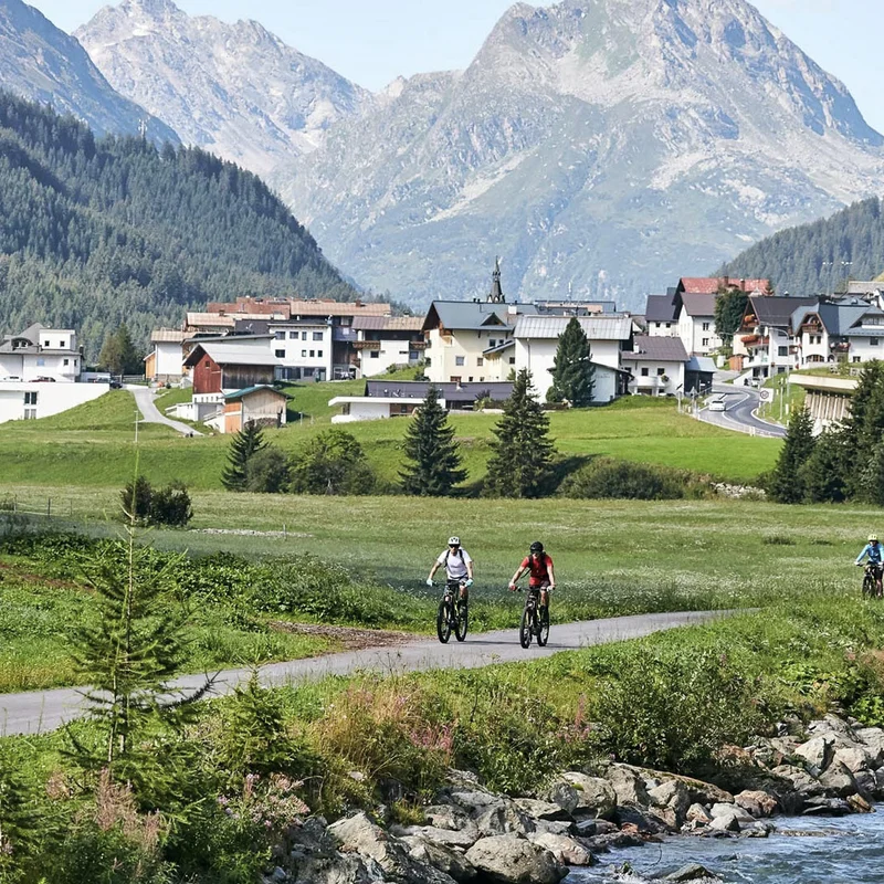 Fahrradfahrer auf Weg entlang Fluss mit Bergdorf und Bergen im Hintergrund