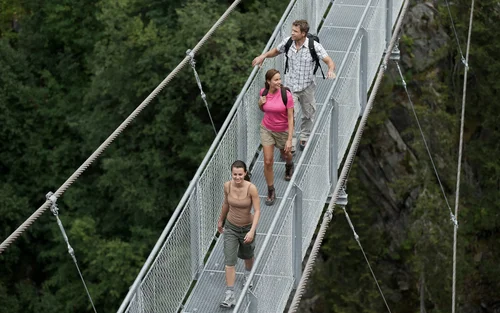 Drei Wanderer gehen auf einer Hängebrücke über einem bewaldeten Tal