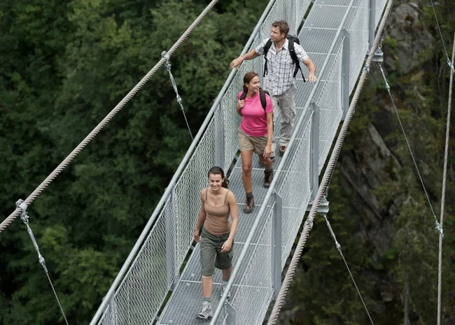 Ihr Sommer im Paznaun im Wander- und Bike-Hotel in Ischgl Drei Wanderer gehen auf einer Hängebrücke über einem bewaldeten Tal