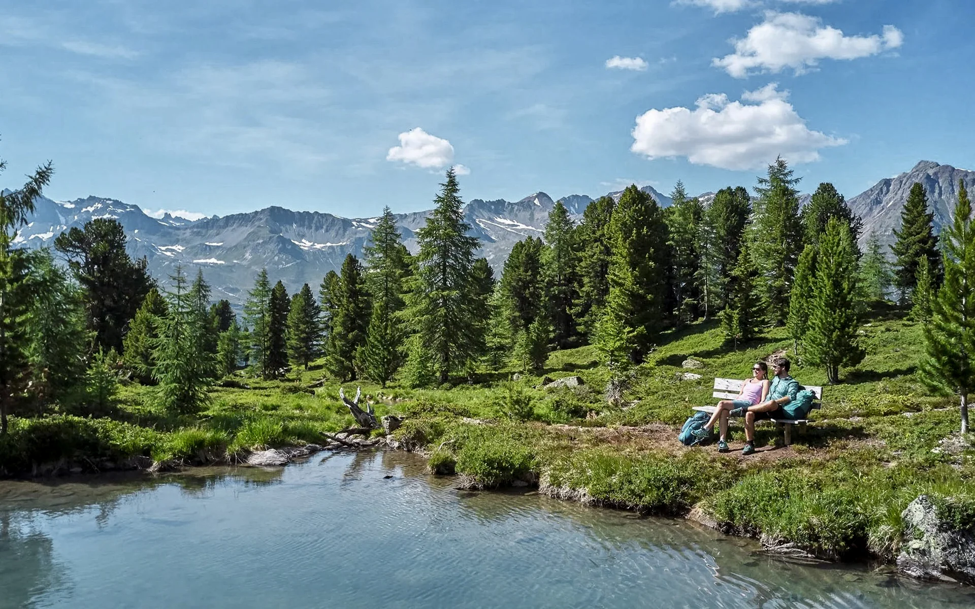 Der Salnerhof – das Hotel in Ischgl Zwei Wanderer sitzen auf einer Bank am See mit Bergpanorama und Tannen