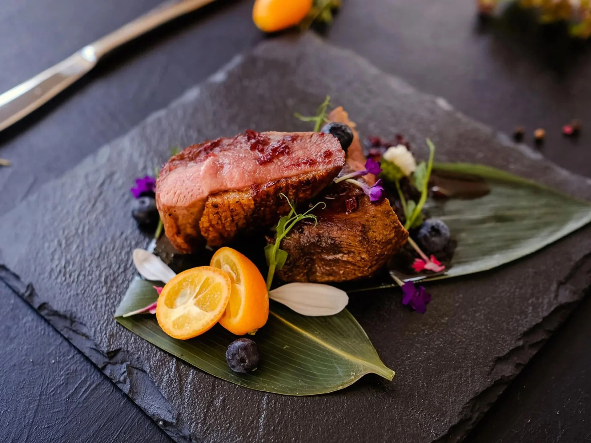Die Bilder aus dem Hotel Salnerhof Gourmetgericht mit Fleisch, Kumquats und essbaren Blüten auf schwarzem Stein