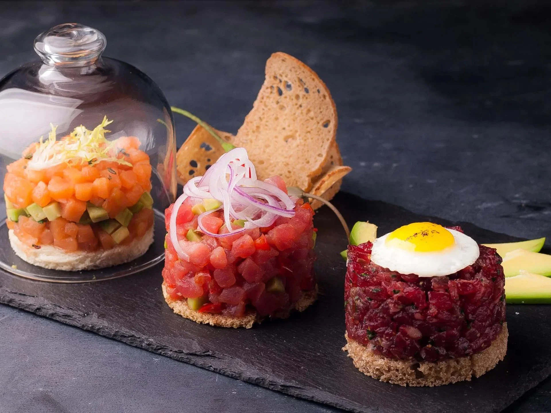Die Bilder aus dem Hotel Salnerhof Drei kunstvoll angerichtete Tartare-Variationen mit Brot und Avocado auf schwarzem Stein