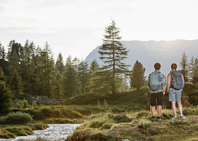 Ihr Sommer im Paznaun im Wander- und Bike-Hotel in Ischgl Zwei Wanderer mit Rucksäcken blicken auf bewaldete Berge im Sonnenlicht