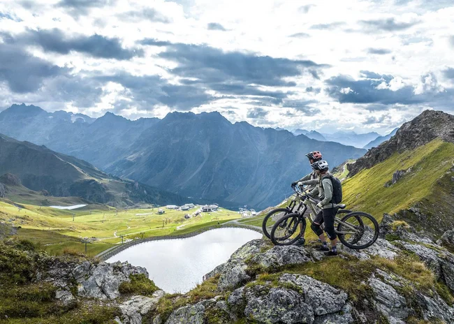 Ihr Sommer im Paznaun im Wander- und Bike-Hotel in Ischgl Zwei Mountainbiker auf einem Felsen mit Blick auf Bergsee und Alpenlandschaft