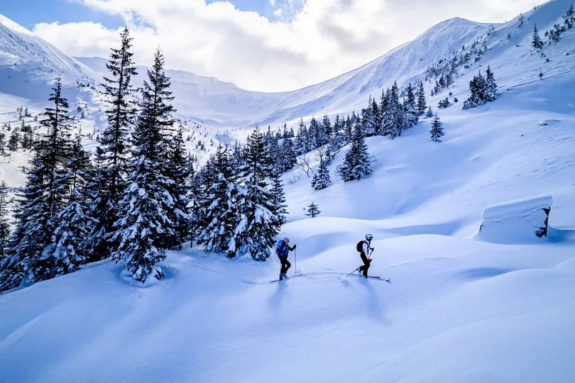 Ihr Skihotel in Ischgl Zwei Skifahrer im verschneiten Bergtal mit Tannenbäumen und blauem Himmel