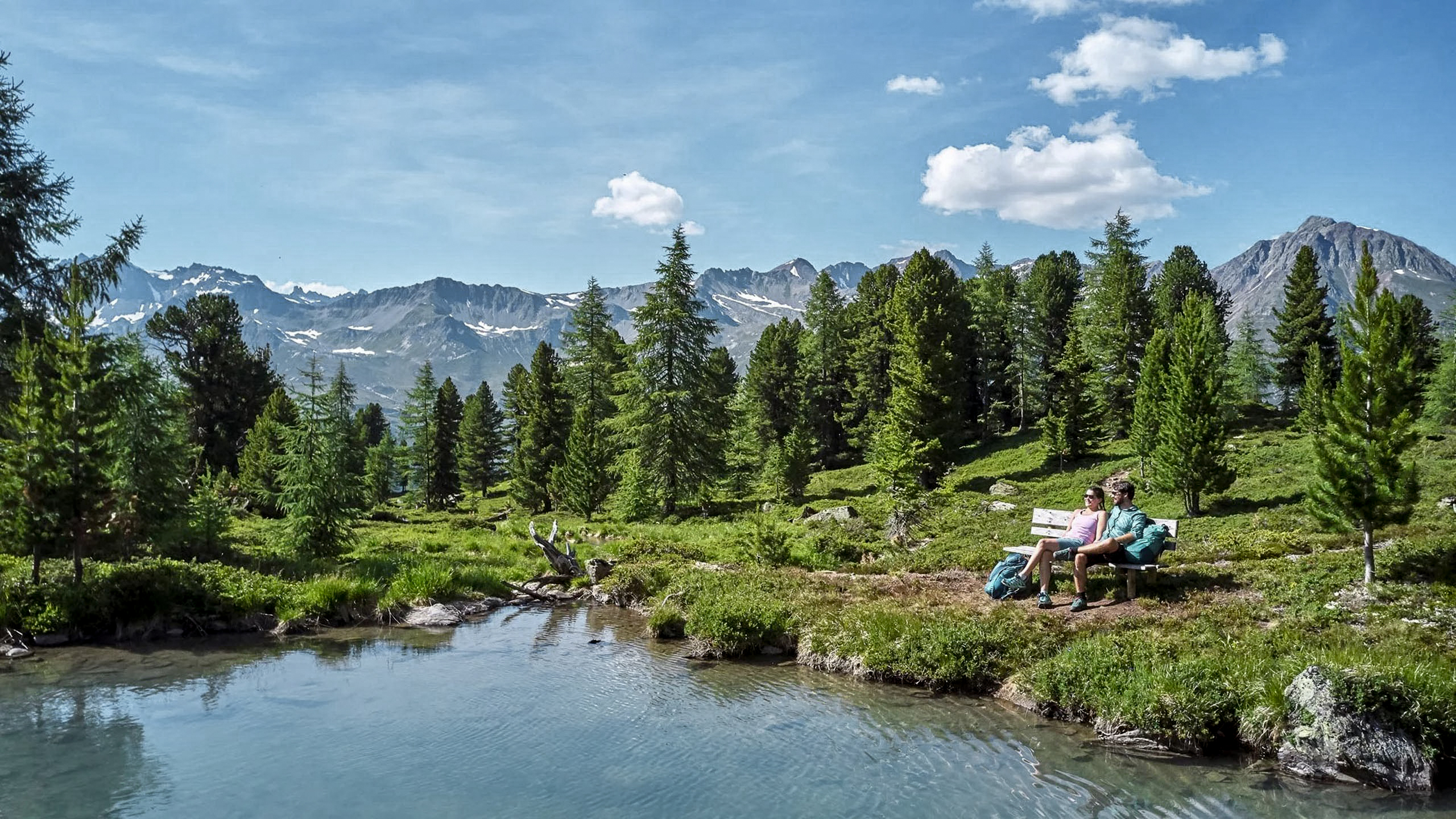 Die Bilder aus dem Hotel Salnerhof Zwei Wanderer sitzen auf einer Bank am See mit Bergpanorama und Tannen