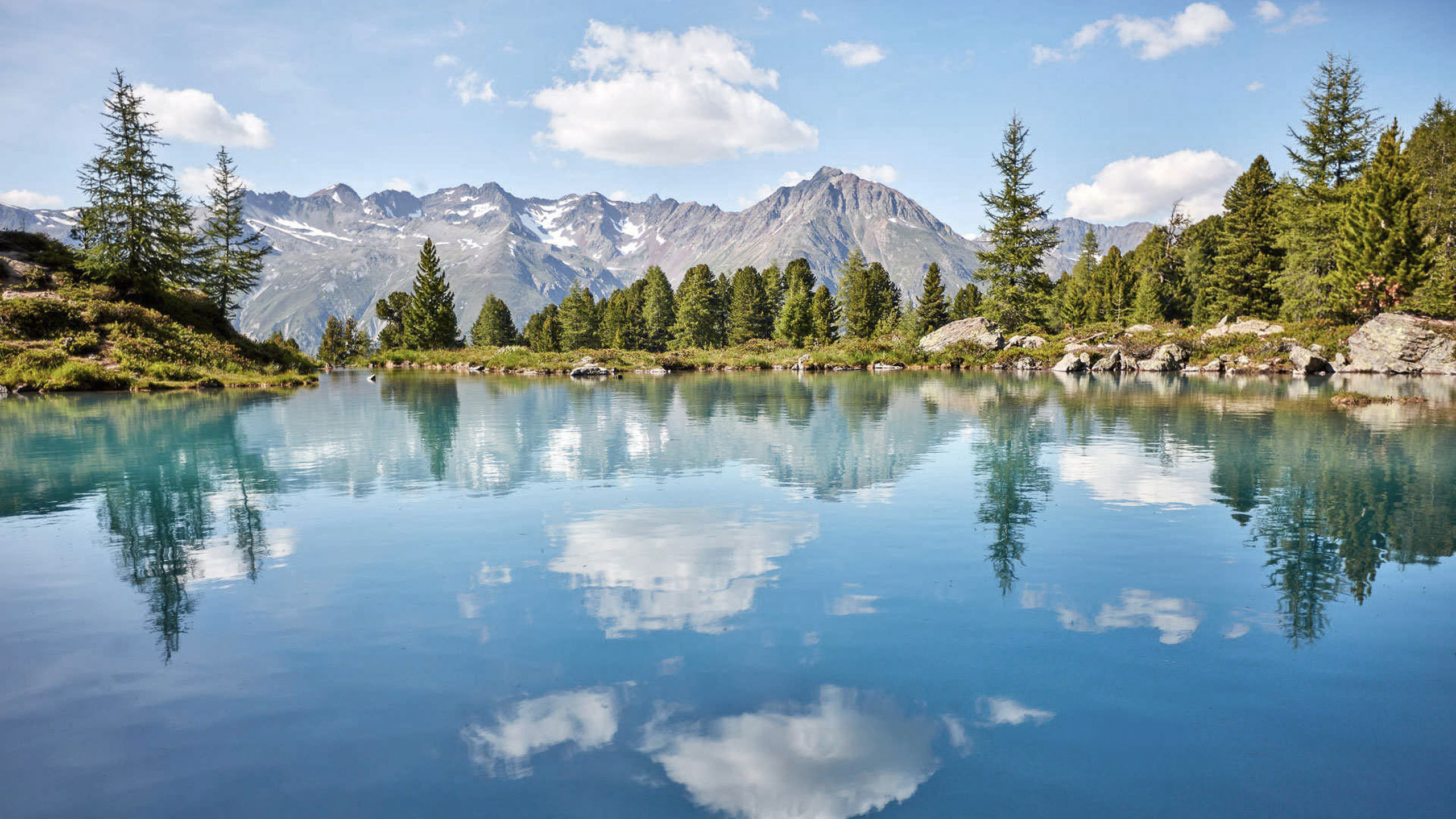 Die Bilder aus dem Hotel Salnerhof Bergsee mit klarer Spiegelung von Bäumen, Bergen und Wolken bei Sonnenschein